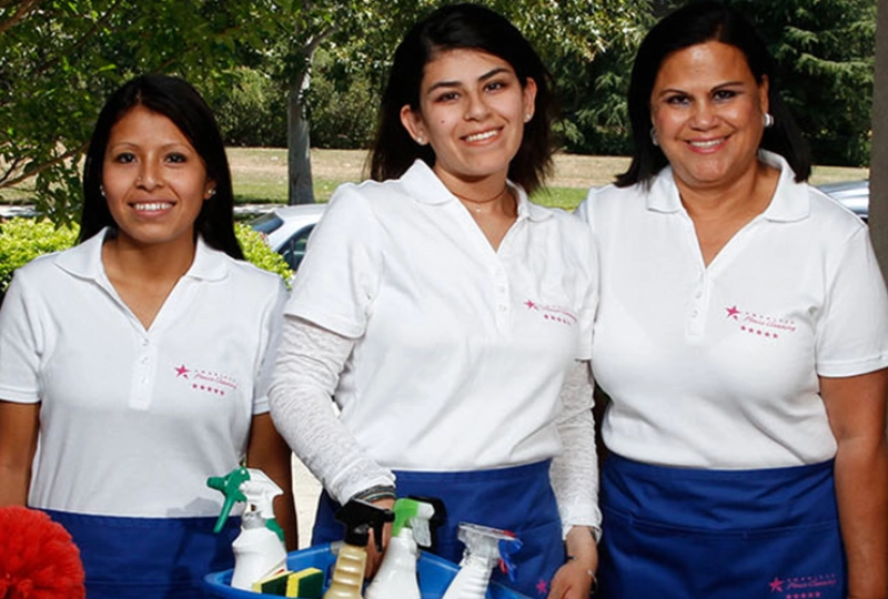 Three women in white shirts and blue aprons holding cleaning supplies.